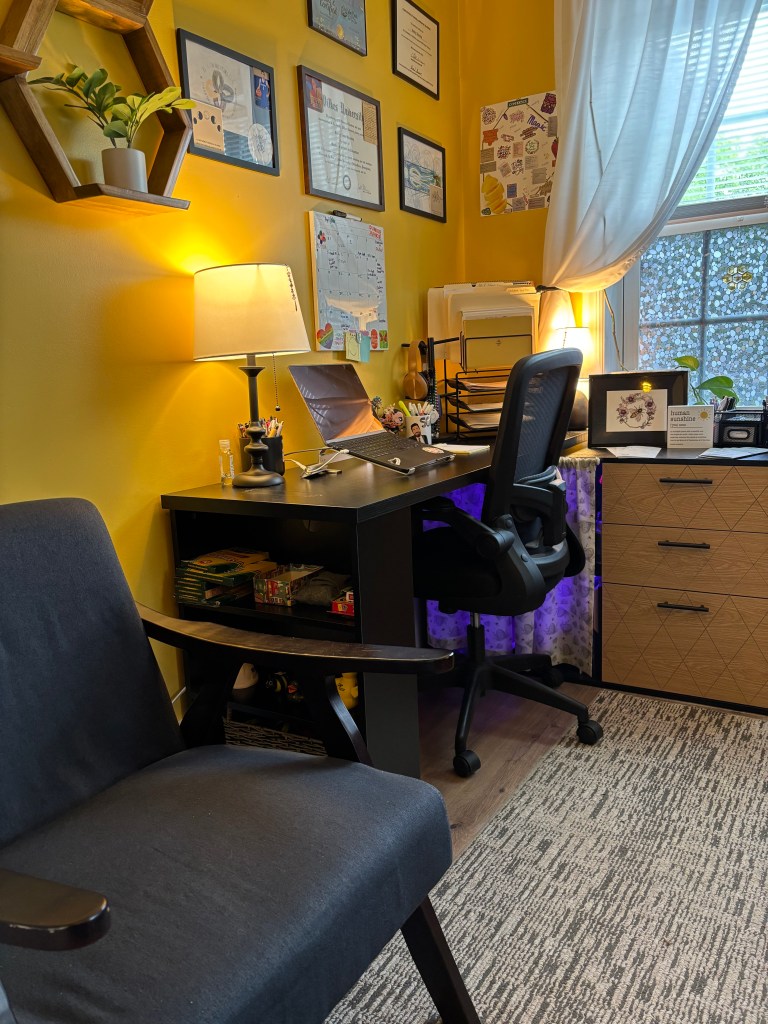 A cozy office space featuring a black desk with a laptop, a comfortable gray chair, and decorated walls with various certificates and a calendar. The room has warm lighting and a window with sheer curtains.